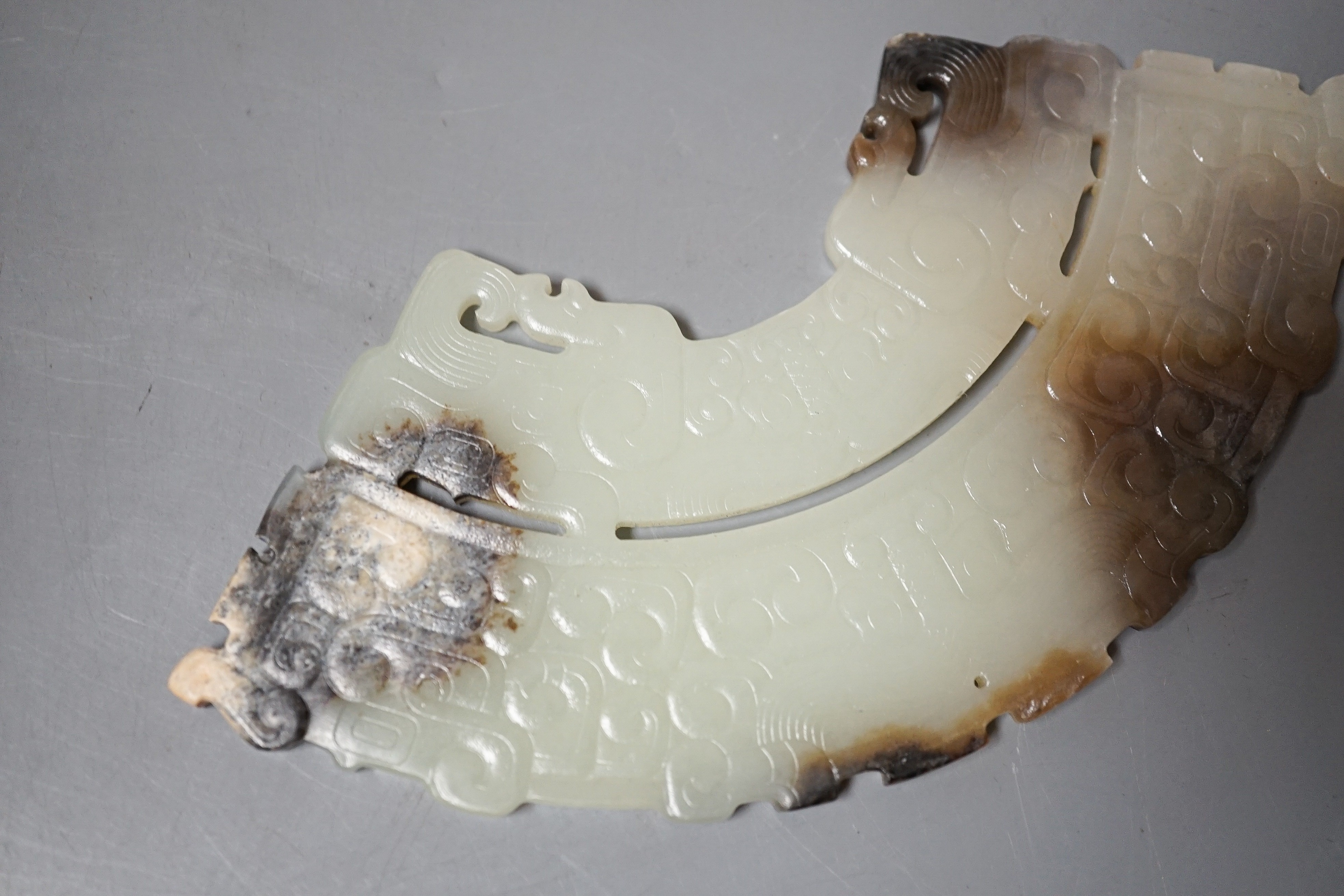 A Chinese archaistic jade fan shaped plaque, the pale celadon stone with brown and black inclusions, carved in low relief with taotie masks and leiwen, width 14.7cm